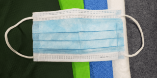 Mask Fabrics (photo): surgical mask on top of a stack of fabrics sometimes used in masks including spunbond nonwoven polypropylene (NWPP), heavy duty surgical wrap (SMS, H600), nanofiber material (Filti), and cotton denim material